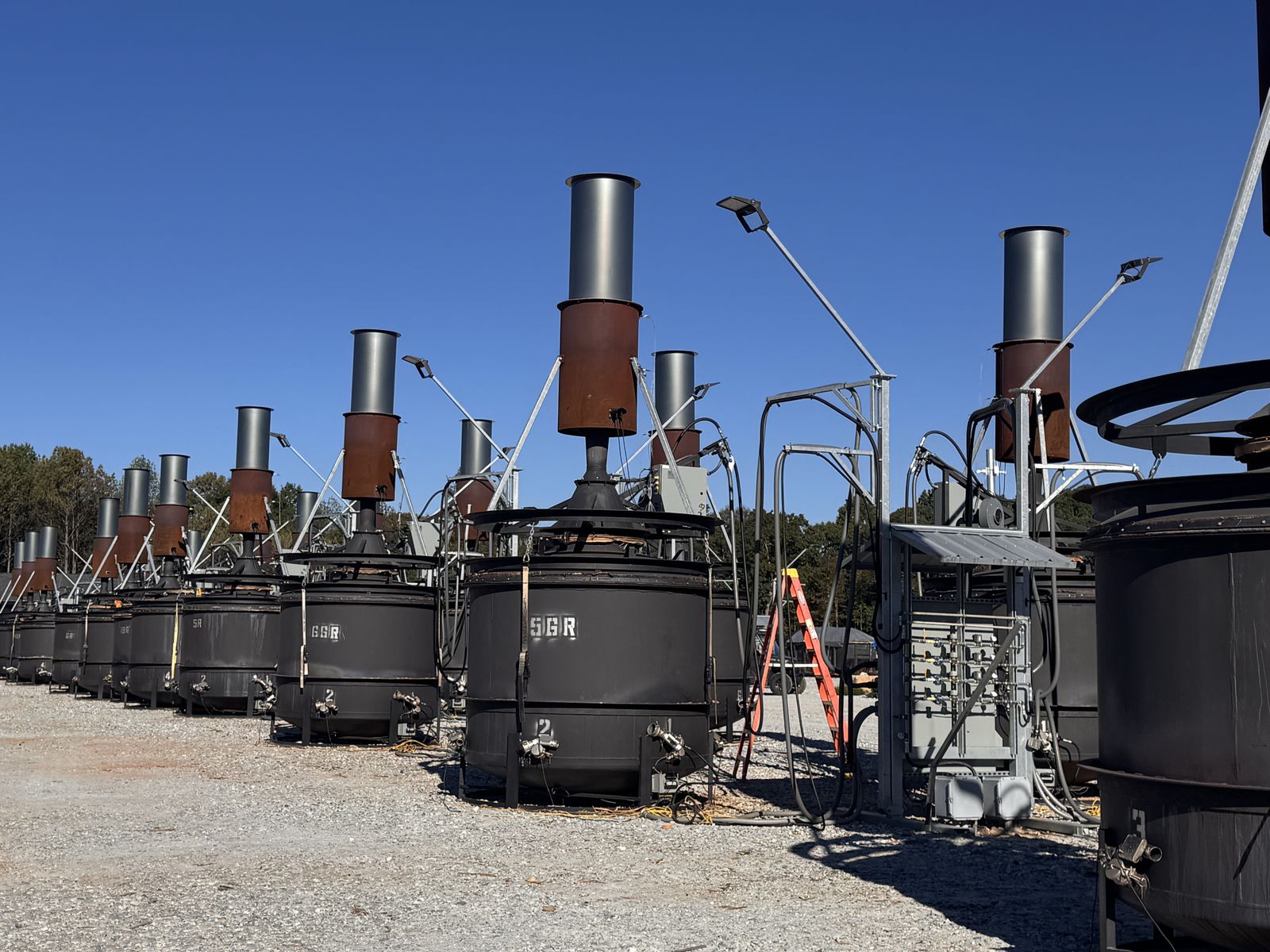 Close view of dark steel kilns with tall metal chimneys, each marked with a number, lined up on a gravel yard.