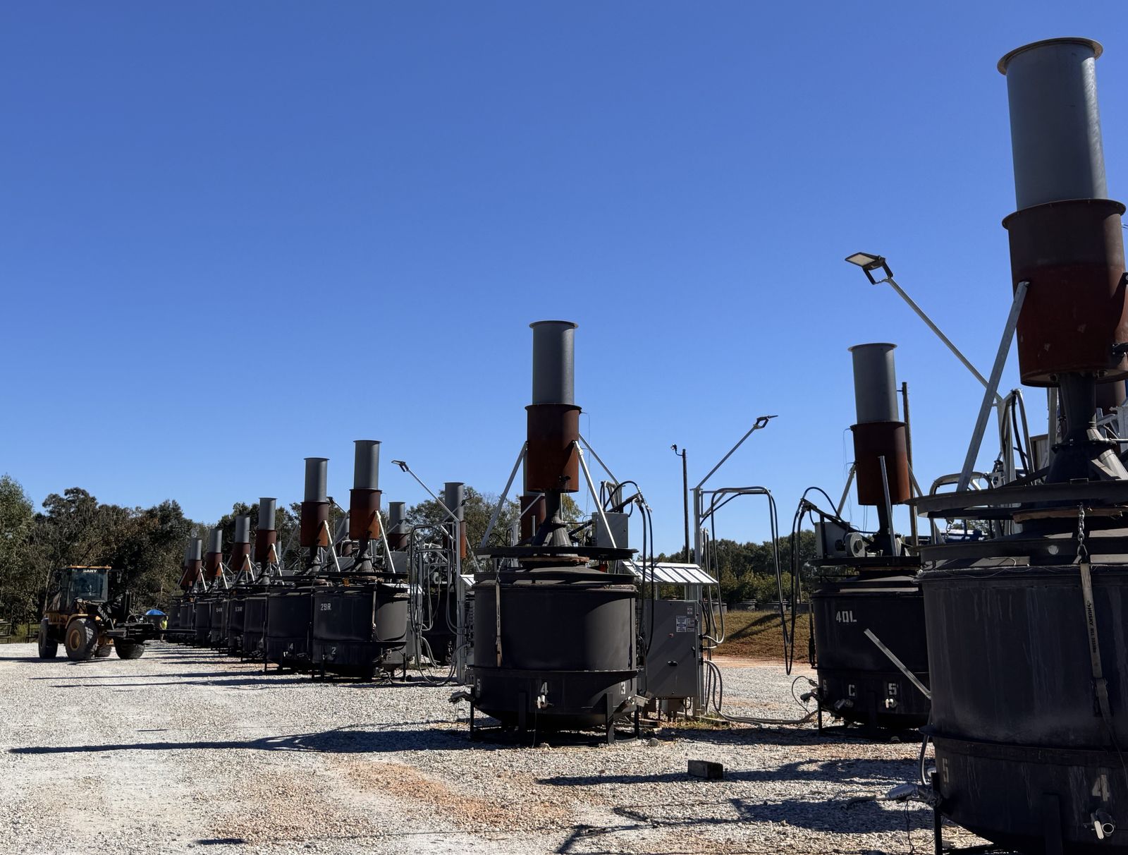 Kilns with tall gray and rust-colored chimneys lined up along a gravel yard, with a wheel loader parked in the distance and trees beyond.