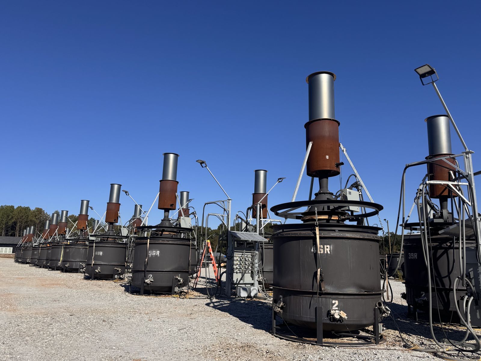 A long row of dark steel kilns, each topped with a tall gray and rust-colored chimney, standing on a gravel yard under a clear blue sky.