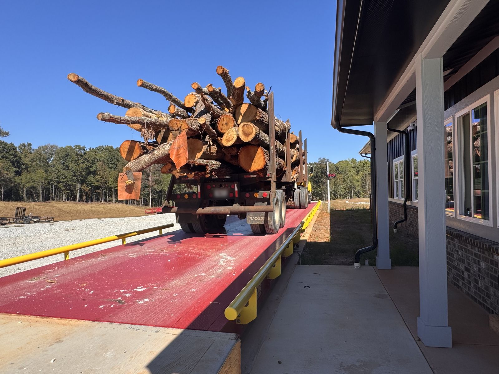 A truck loaded with cut logs sits on a red truck scale with yellow safety rails, beside the scale house.