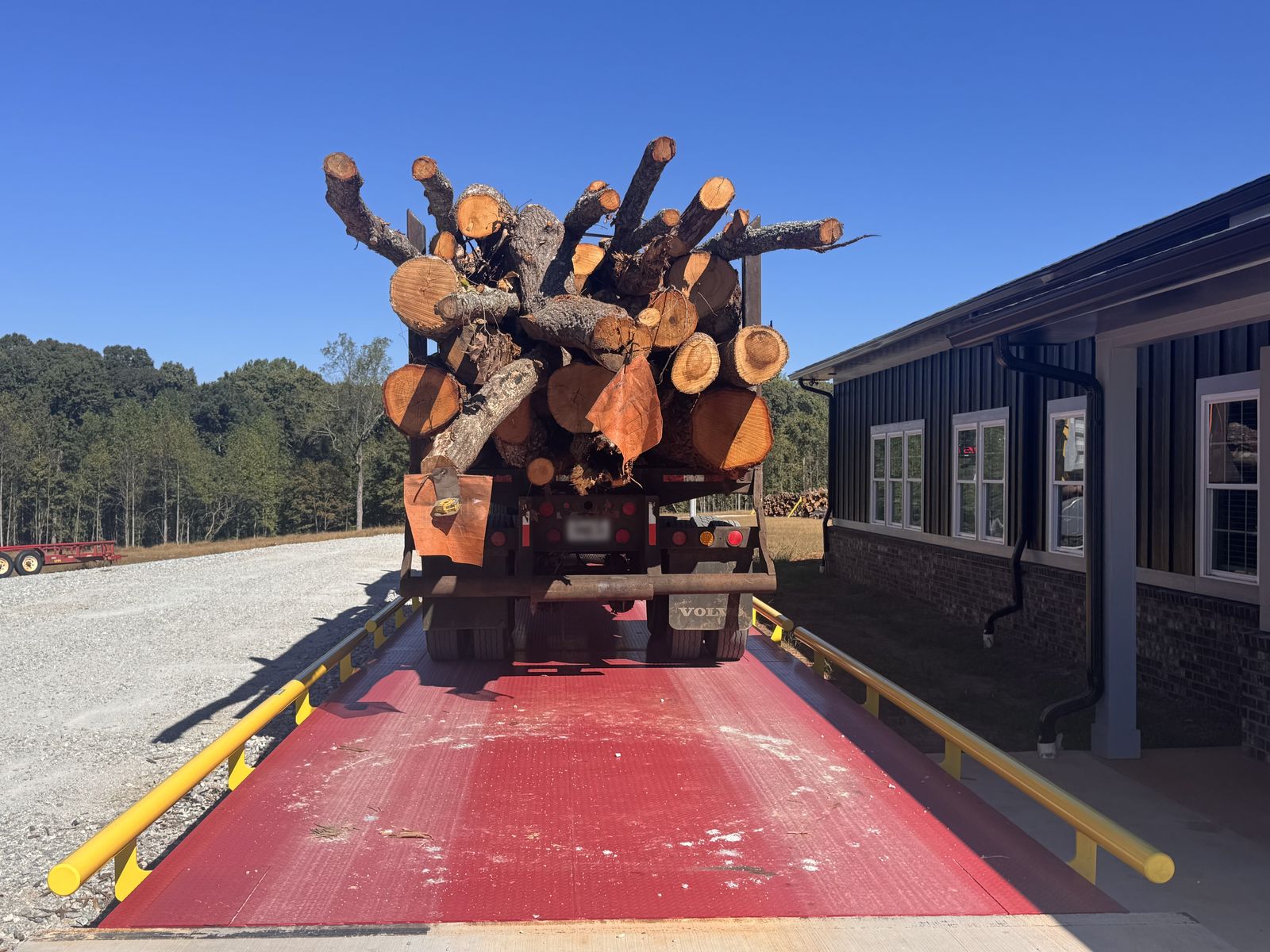 A truck loaded with logs, seen from behind, stopped on a red truck scale with yellow rails beside the scale house.